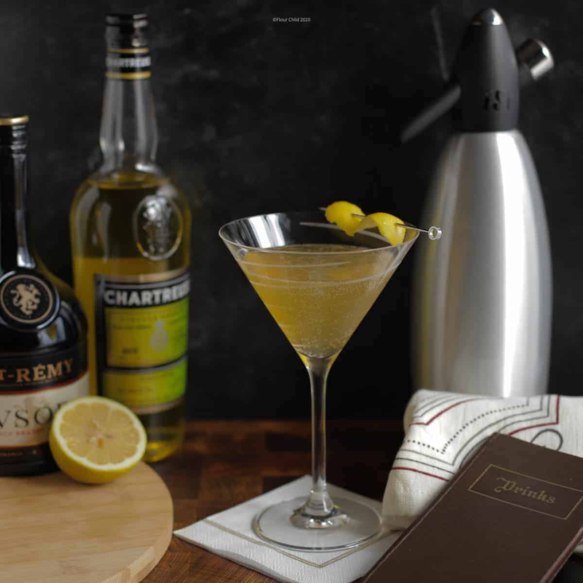 A Brandy Daisy cocktail shown with a lemon twist in a cocktail glass surrounded by its ingredients