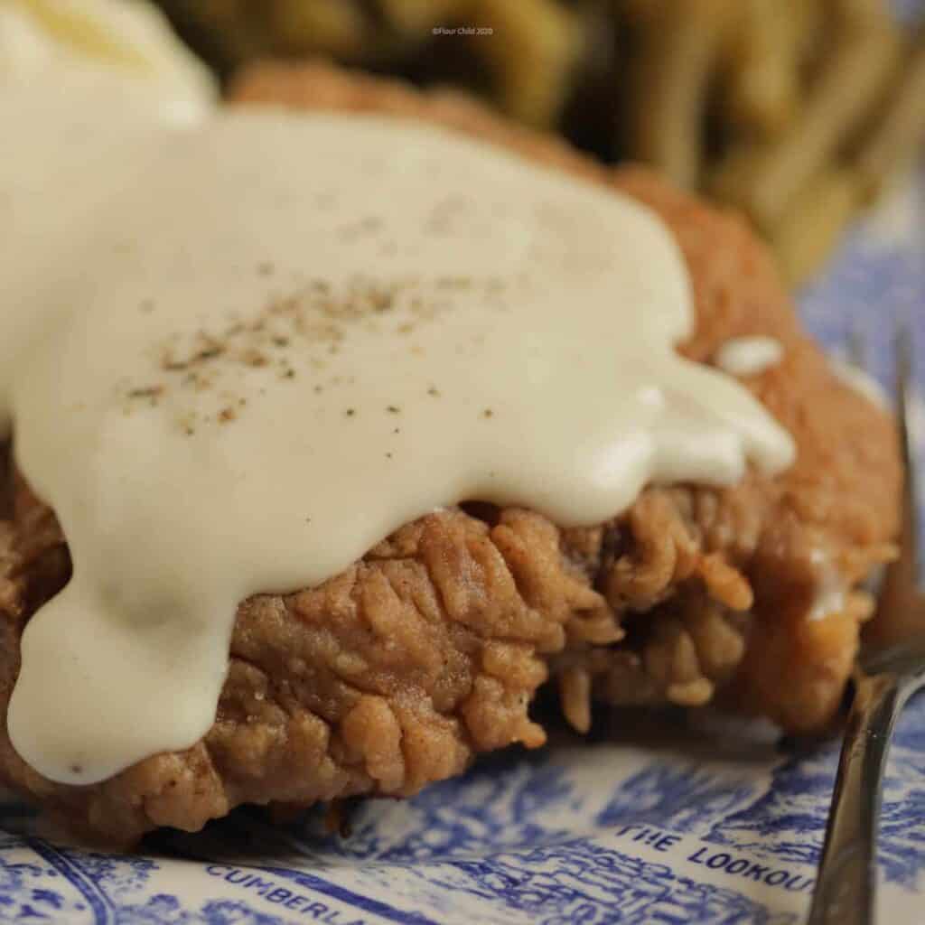 A chicken fried steak on a dinner plate covered with white pepper gravy, sitting next to green beans and mashed potatoes
