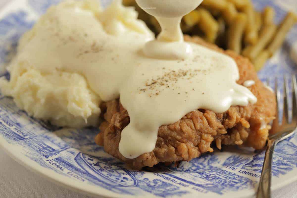 Chicken Fried Steak on a plate next to mashed potatoes and green beans, with white gravy being poured over steak.