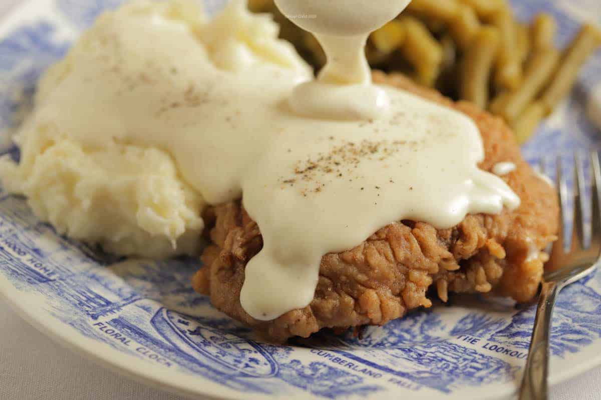 A close up of chicken fried steak on a plate with white pepper gravy being poured over the top