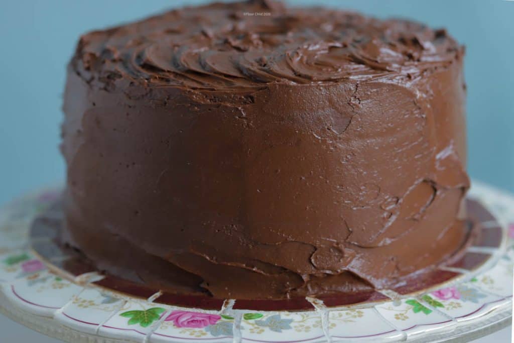 Chocolate cake covered with chocolate buttercream frosting sitting on a round cake platter