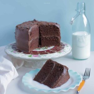 A cake on a pedestal next to a jug of milk, with a slice of cake on a plate in front of it