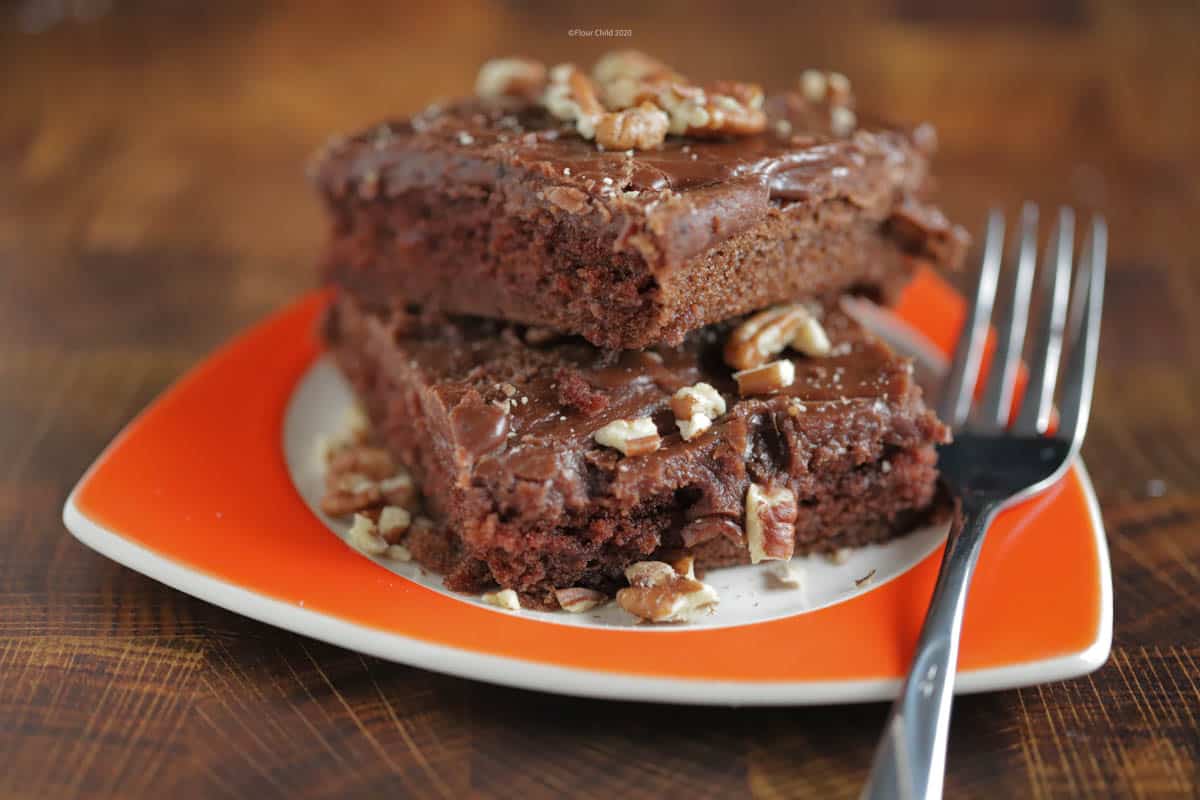 Two brownies with nuts on top sitting on an orange plate with a fork next to it