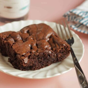 A square brownie on an ivory plate with a glass of milk above it and a fork next to it
