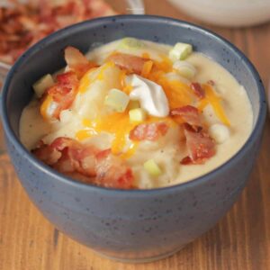 A bowl of baked potato soup topped with bacon, green onions, shredded cheddar, and a dollop of sour cream