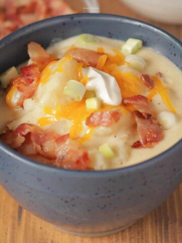 A bowl of baked potato soup topped with bacon, green onions, shredded cheddar, and a dollop of sour cream