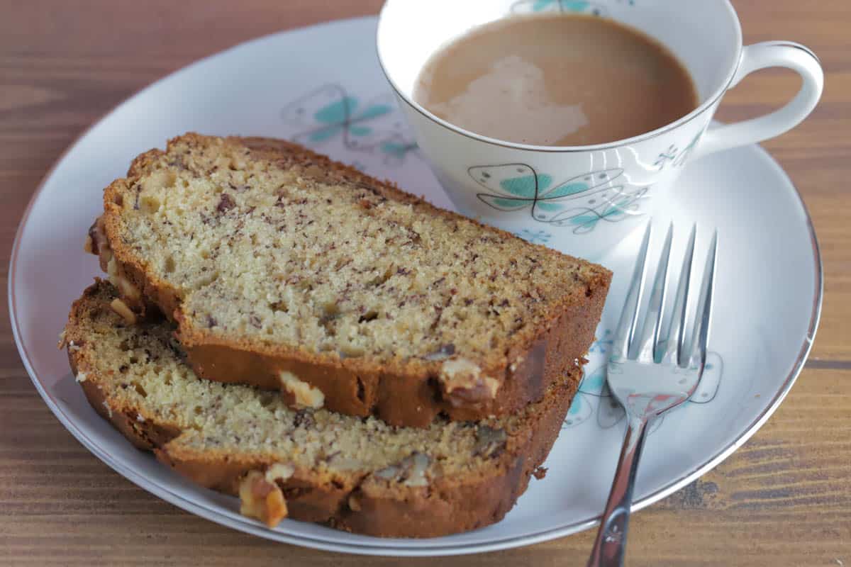 Two slices of banana walnut bread on a plate, with a fork and a cup of coffee next to it.