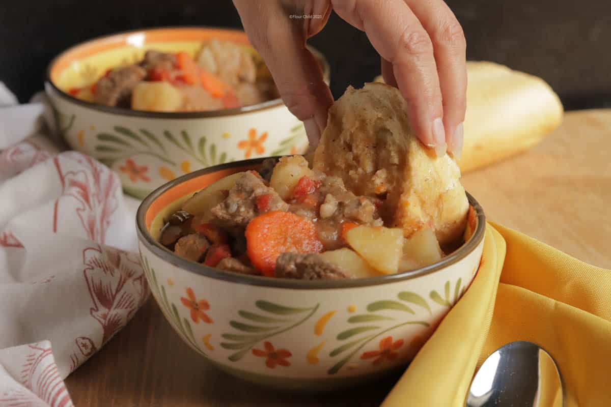 A bowl of beef burgundy stew with chunks of meat, potatoes, and vegetables piled high. A hand is dipping a piece of French bread into the stew.