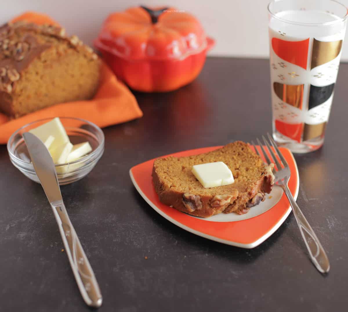 A slice of pumpkin bread with a pat of butter on top, sitting on an orange plate next to a glass of milk. 