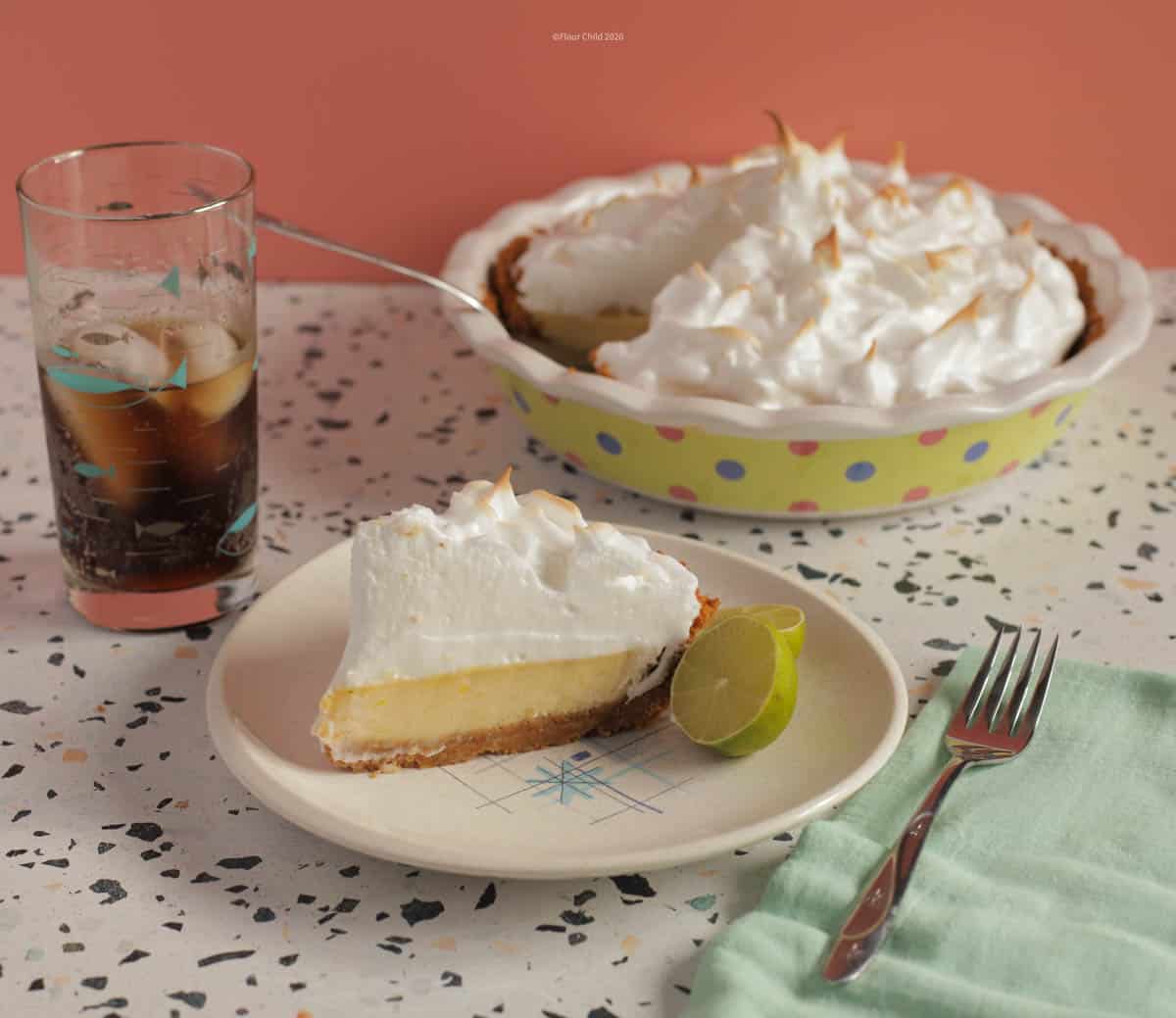 A lice of key lime pie on a plate with a lime wedge next to it. Pie dish in the background and a glass of coke and a fork next to the plate.