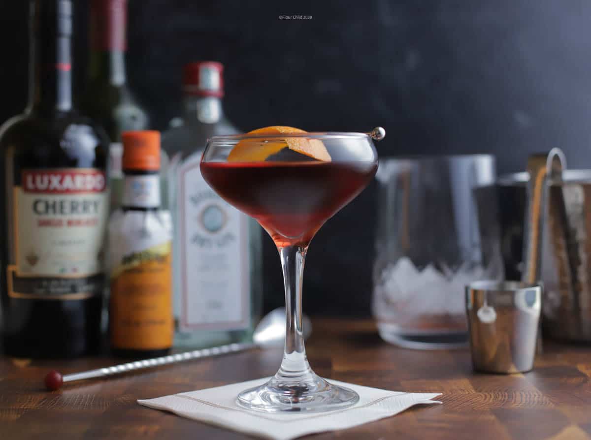 A Martinez cocktail in a coupe glass surrounded by ingredients bottles and a mixing glass