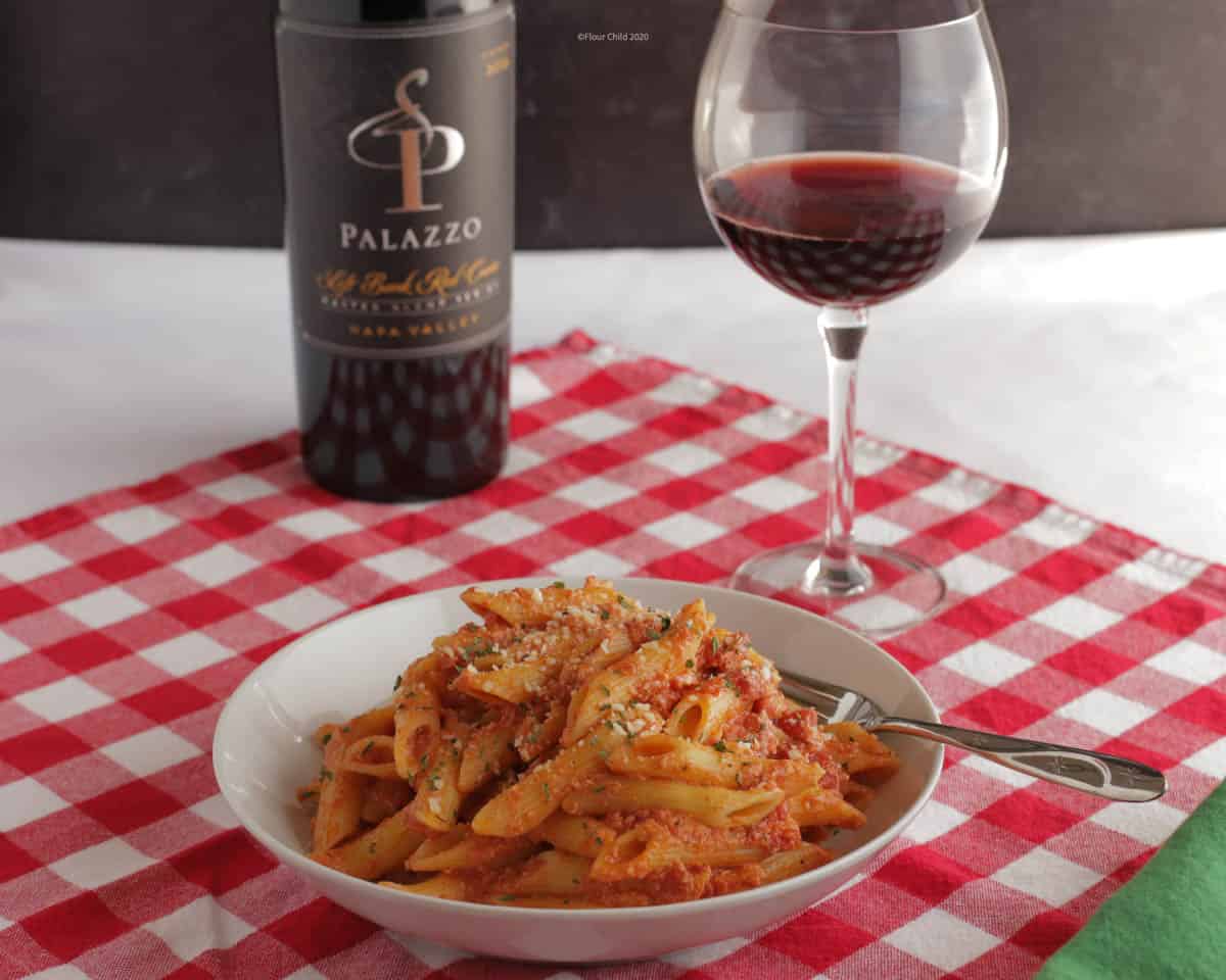 Penne pasta in vodka sauce in a white bowl with a fork, a bottle of wine and a glass of wine in the background