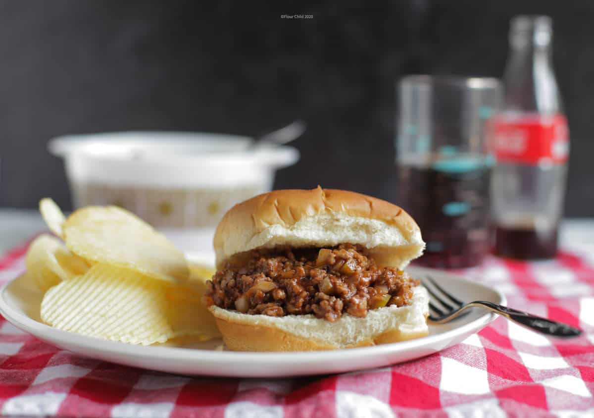 Sloppy Joe on a bun, sitting on a plate with potato chips and a fork