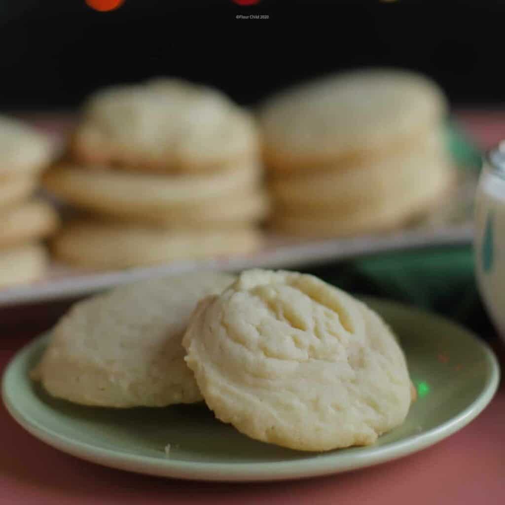 Two butter cookies on a green plate, with a tray of butter cookies behind it