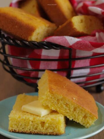 A piece of cornbread sliced in half on a small blue bread plate, with a pat of butter on one piece. A basket of cornbread is in the background.