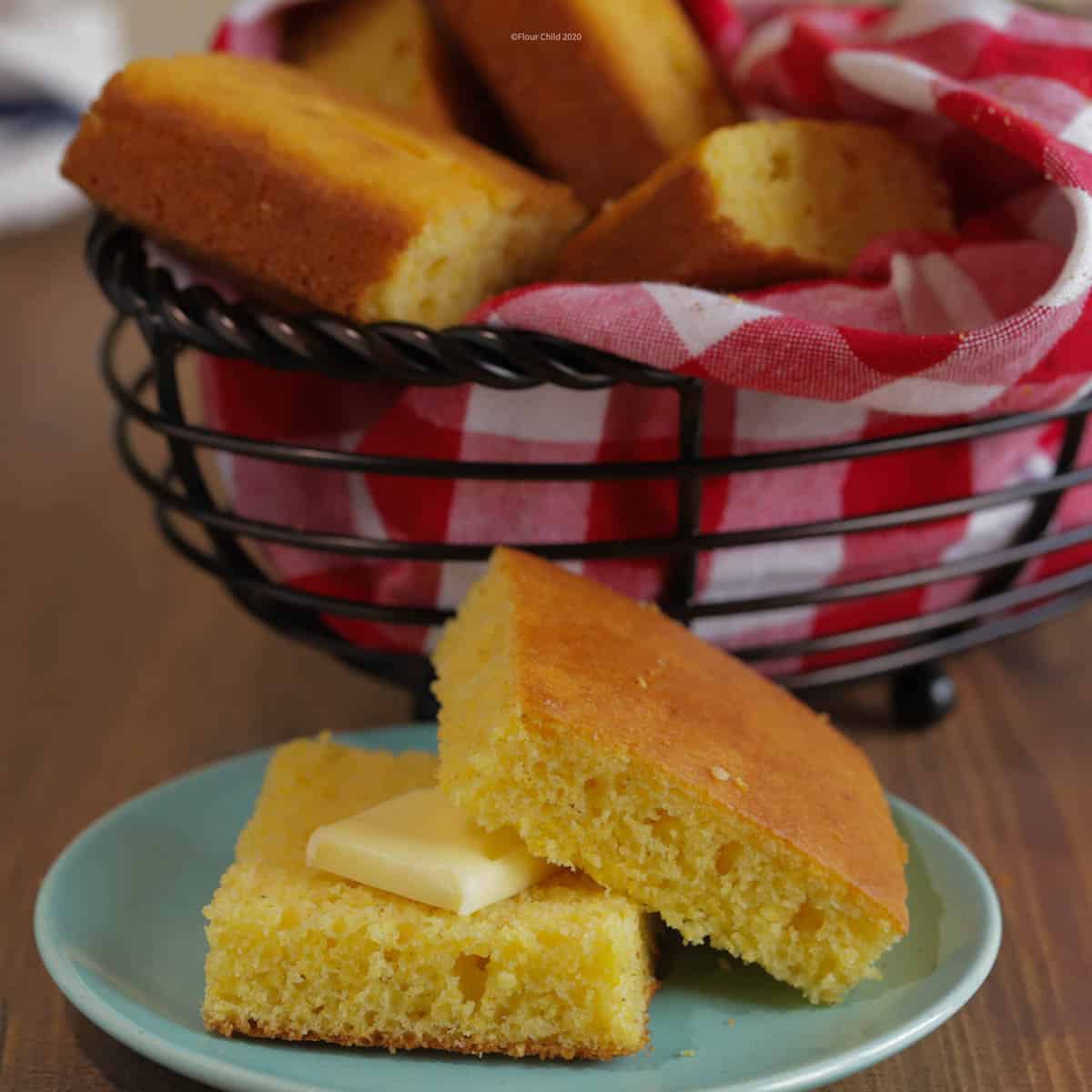 A piece of cornbread sliced in half on a small blue bread plate, with a pat of butter on one piece. A basket of cornbread is in the background.