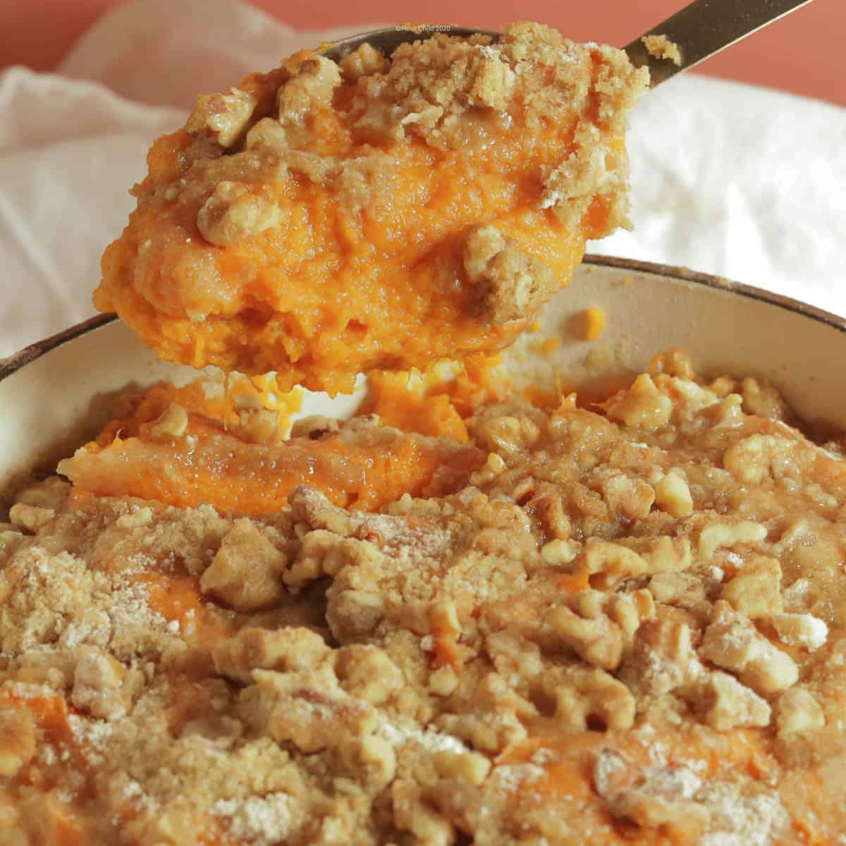 sweet potato casserole with pecan topping in a white cast iron casserole dish, with a large spoon lifting out a serving.