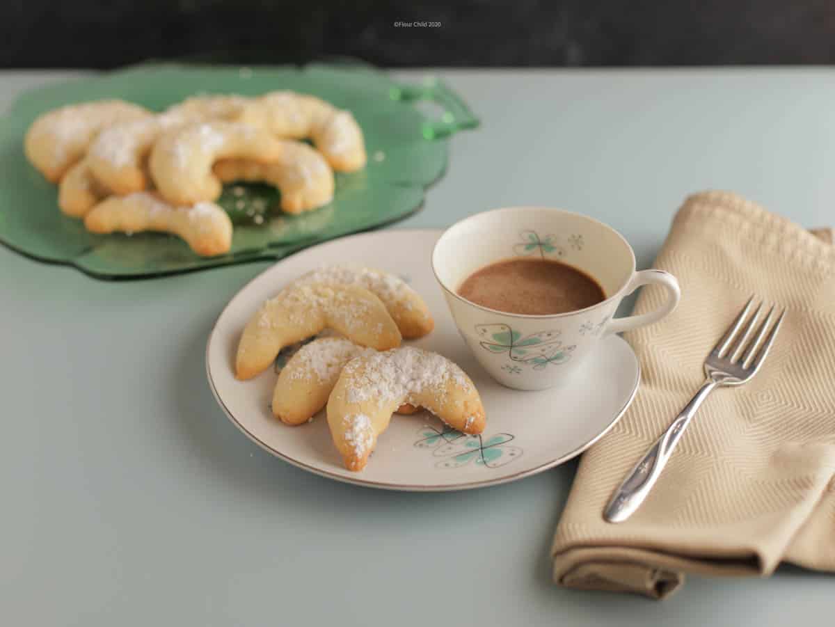 A plate with 3 crescent cookies on it next to a cup of coffee, with a serving tray of cookies in th background