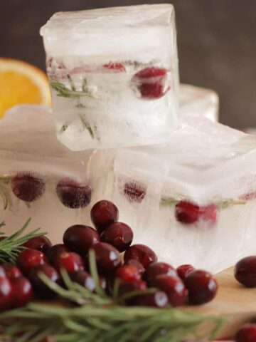 3 giant holiday ice cubes filled with cranberries and rosemary are stacked on a wooden cutting board
