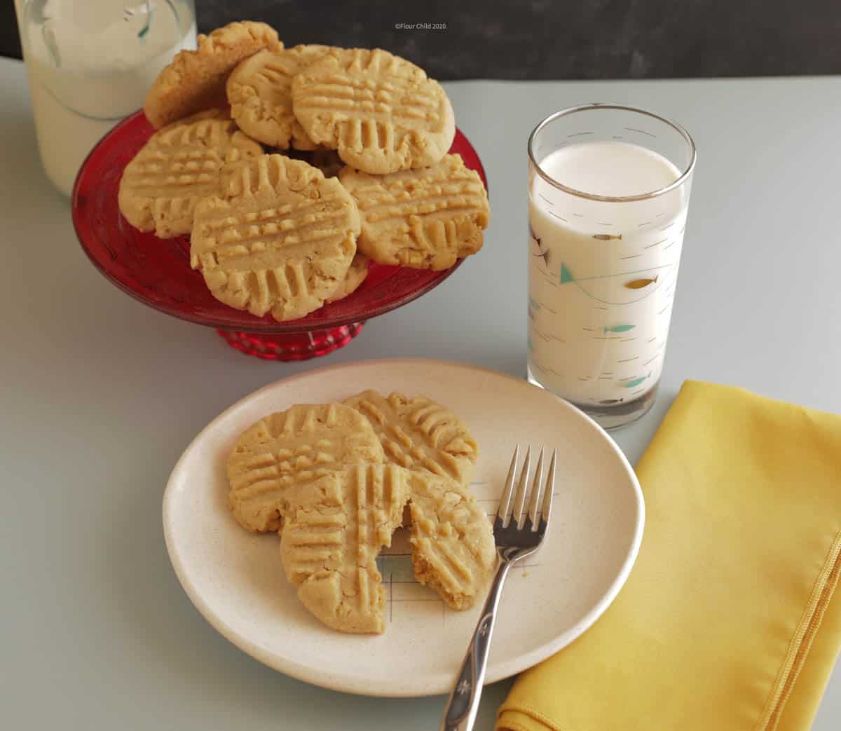 Peanut butter cookies on a dessert plate with a glass of milk next to it and a platter of peanut butter cookies behind it