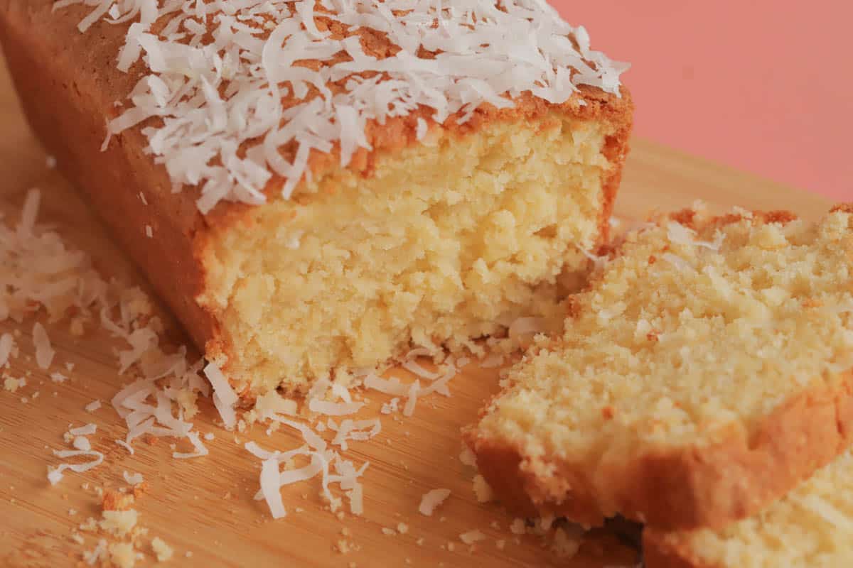 A slice of coconut poundcake in front of the whole poundcake, with coconut sprinkled over the top