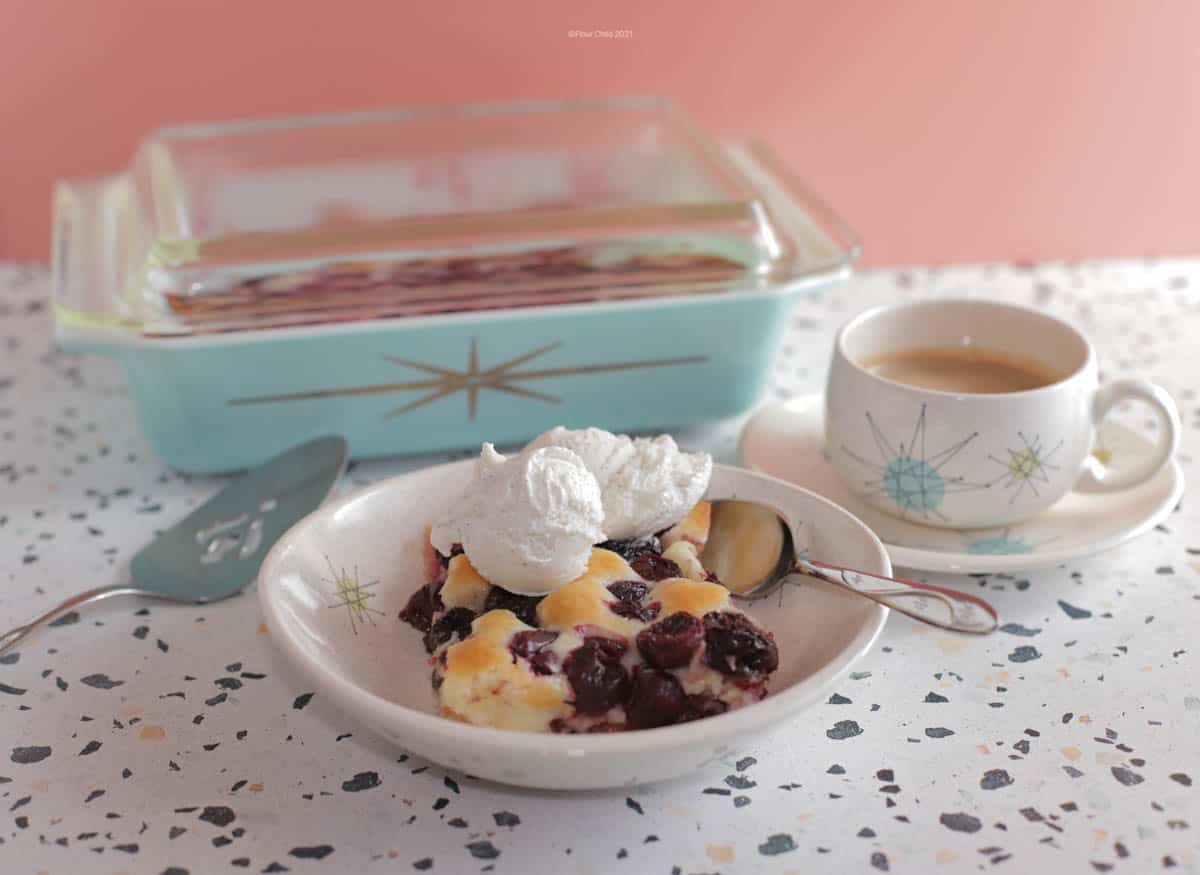 Cherry cobbler in a bowl with ice cream on top, coffee cup and casserole dish in background