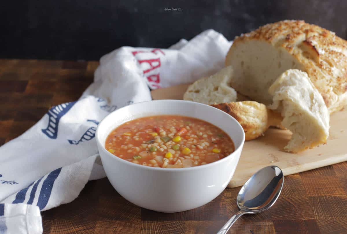 Vegetable barley soup on a bowl with a loaf of bread in the background