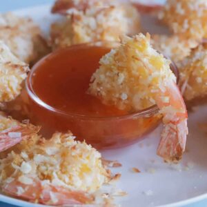 Coconut shrimp on a plate, with a small bowl of apricot dipping sauce that has a shrimp dipped into it.