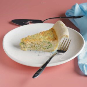 A slice of broccoli & cheddar jack cheese quiche on a plate with a fork next to it.