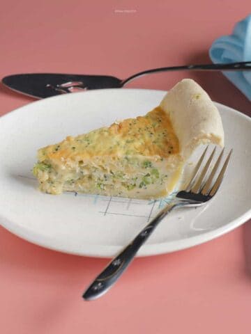 A slice of broccoli & cheddar jack cheese quiche on a plate with a fork next to it.