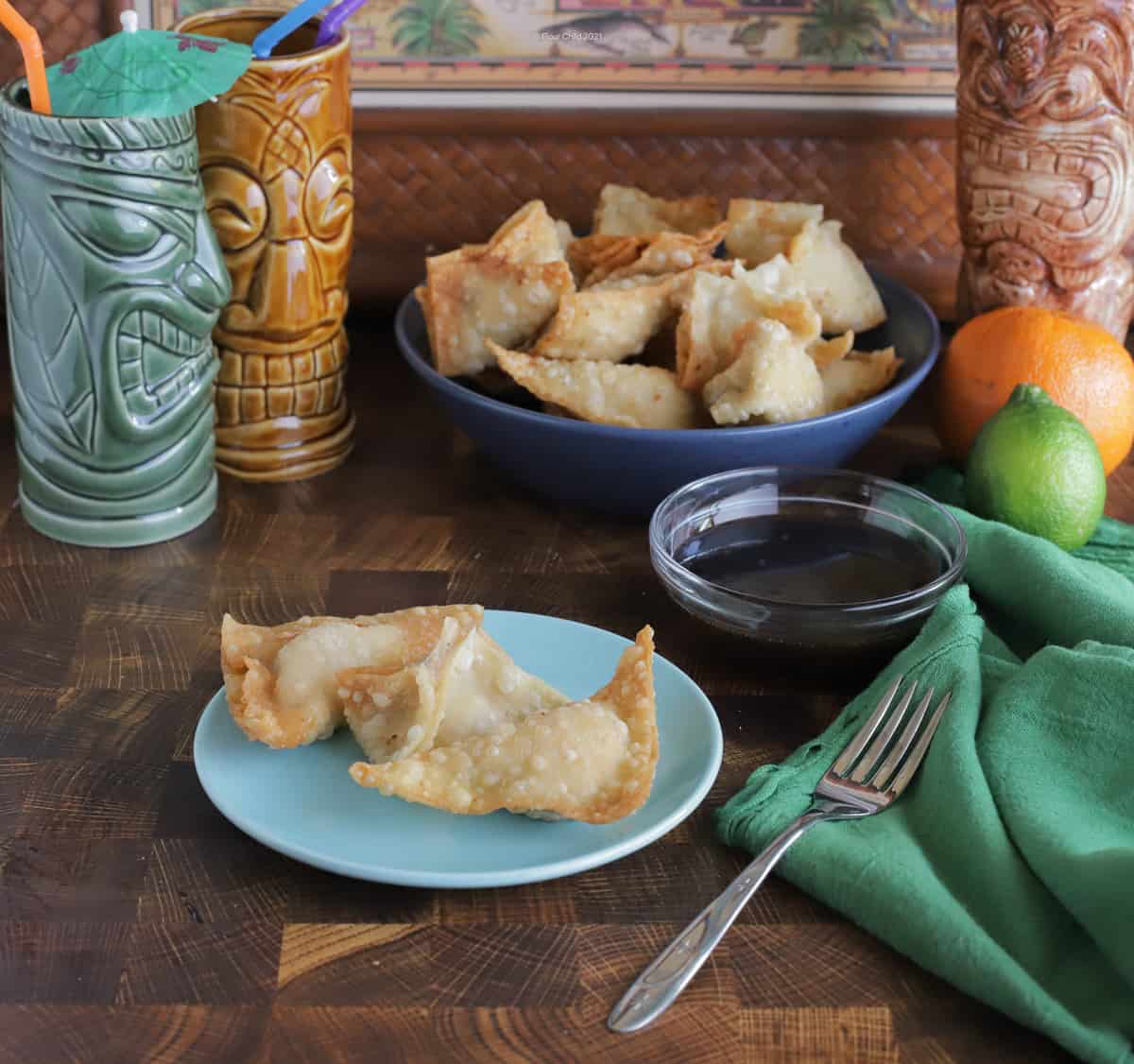 Two pieces of crab rangoon on a blue plate with a blue bowl of crab rangoon in the background