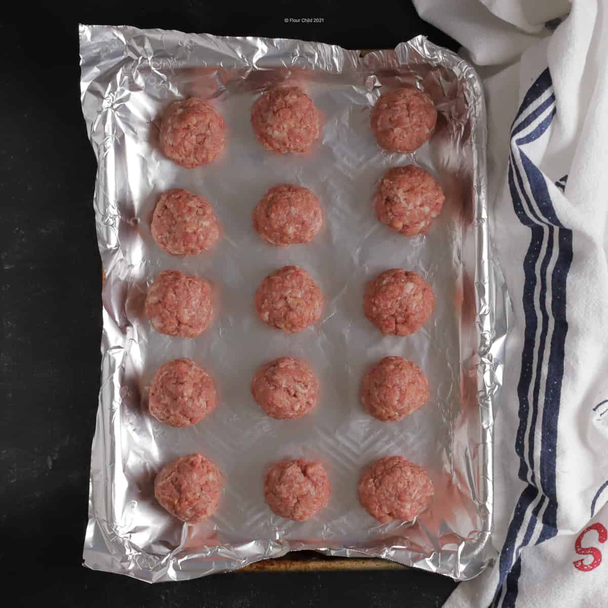 Fifteen uncooked meatballs on a foil lined cookie sheet, ready for baking