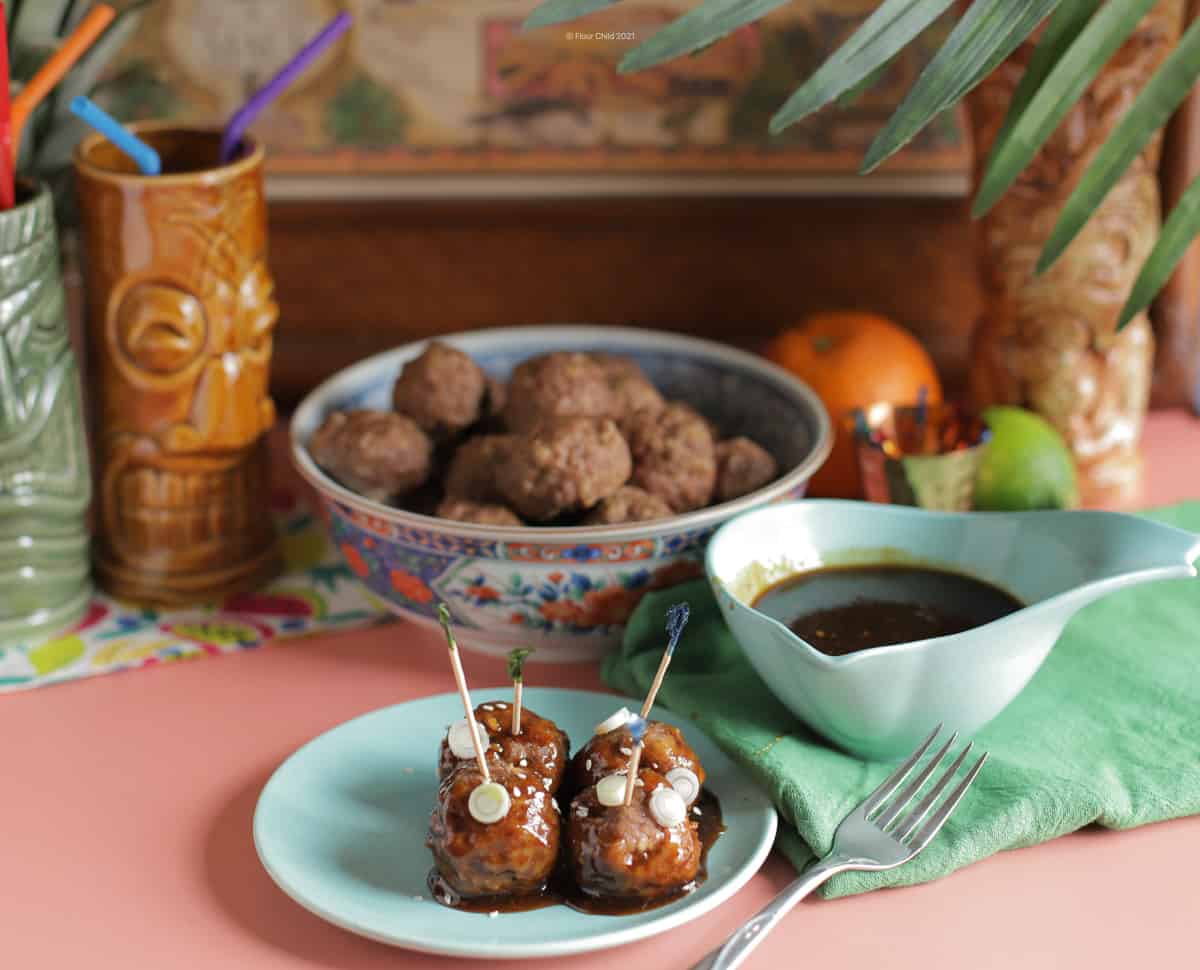 Four meatballs on a small plate with a side of teriyaki sauce, and a bowl of additional meatballs in the background