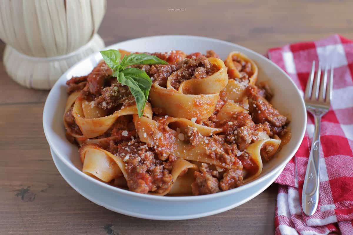 A white bowl filled with homemade bolognese sauce with pappardelle pasta and topped with a basil leaf