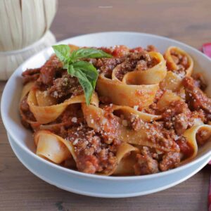 homemade bolognese sauce with pepperdelle pasta with a basil leaf on top in a white bowl.