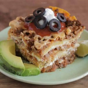 Slice of Mexican Lasagna on a green plate topped with sour cream and olives, with sliced avocados and lime on the side.