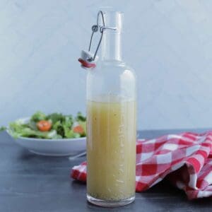 A bottle of homemade vinegar and oil salad dressing on a table, with a red checked napkin on the table and a green salad in the background.