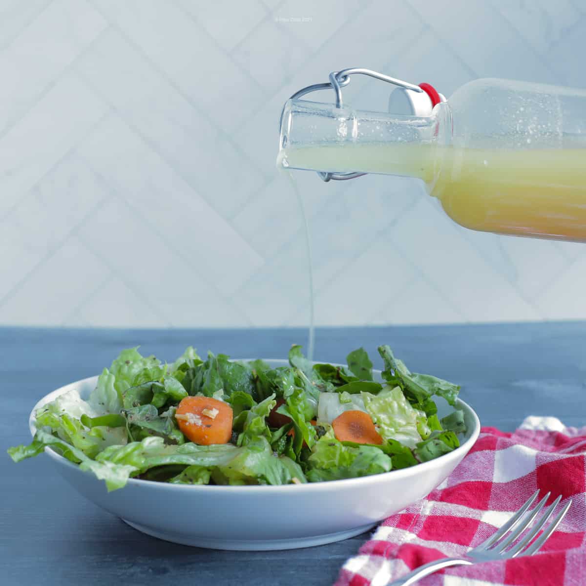 Salad dressing in a glass bottle, being poured over a green salad I a white bowl, with a red checked napkin next to it