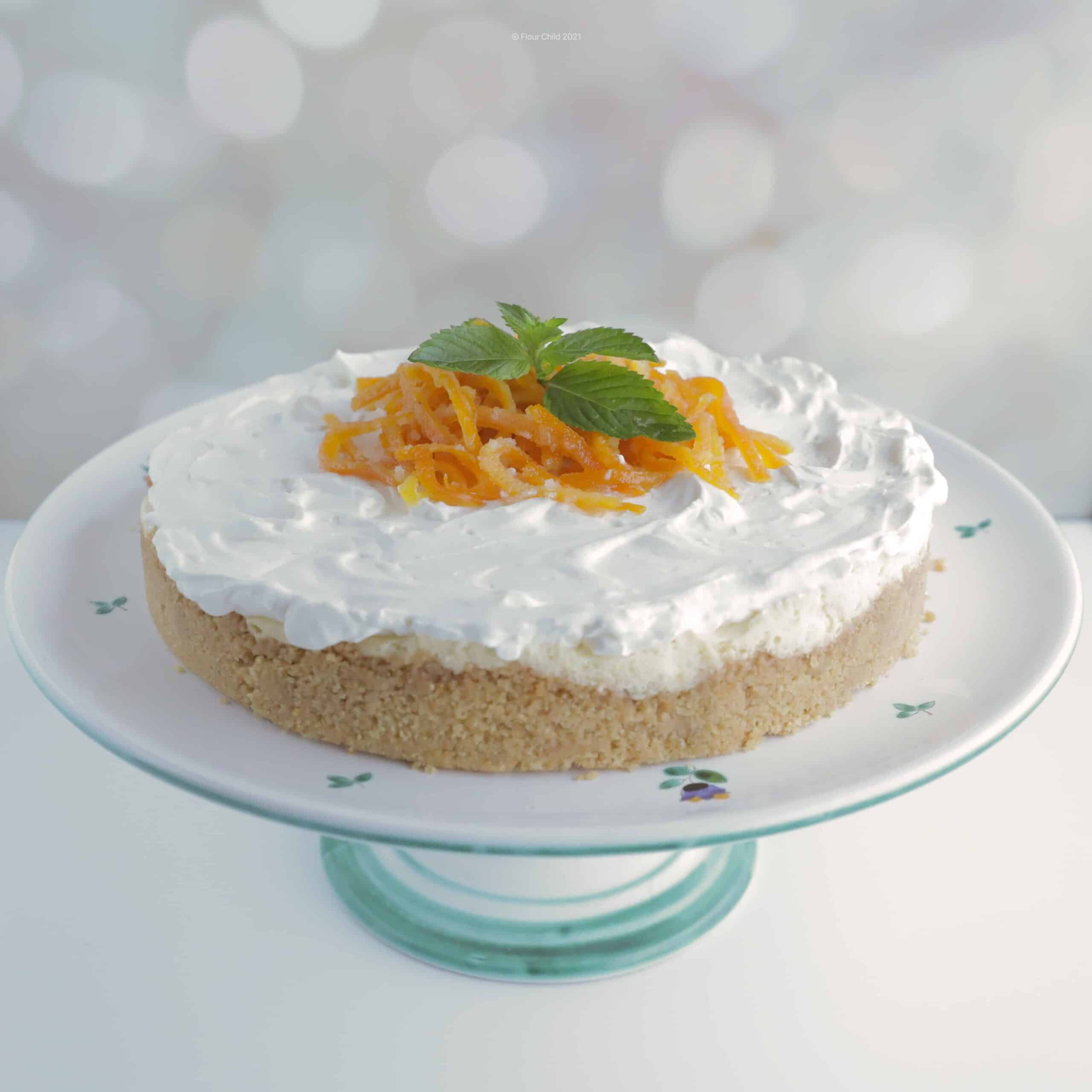 Orange creamsicle cheesecake on a cake pedestal with candied orange peel and mint leaves garnishing the top in the center.