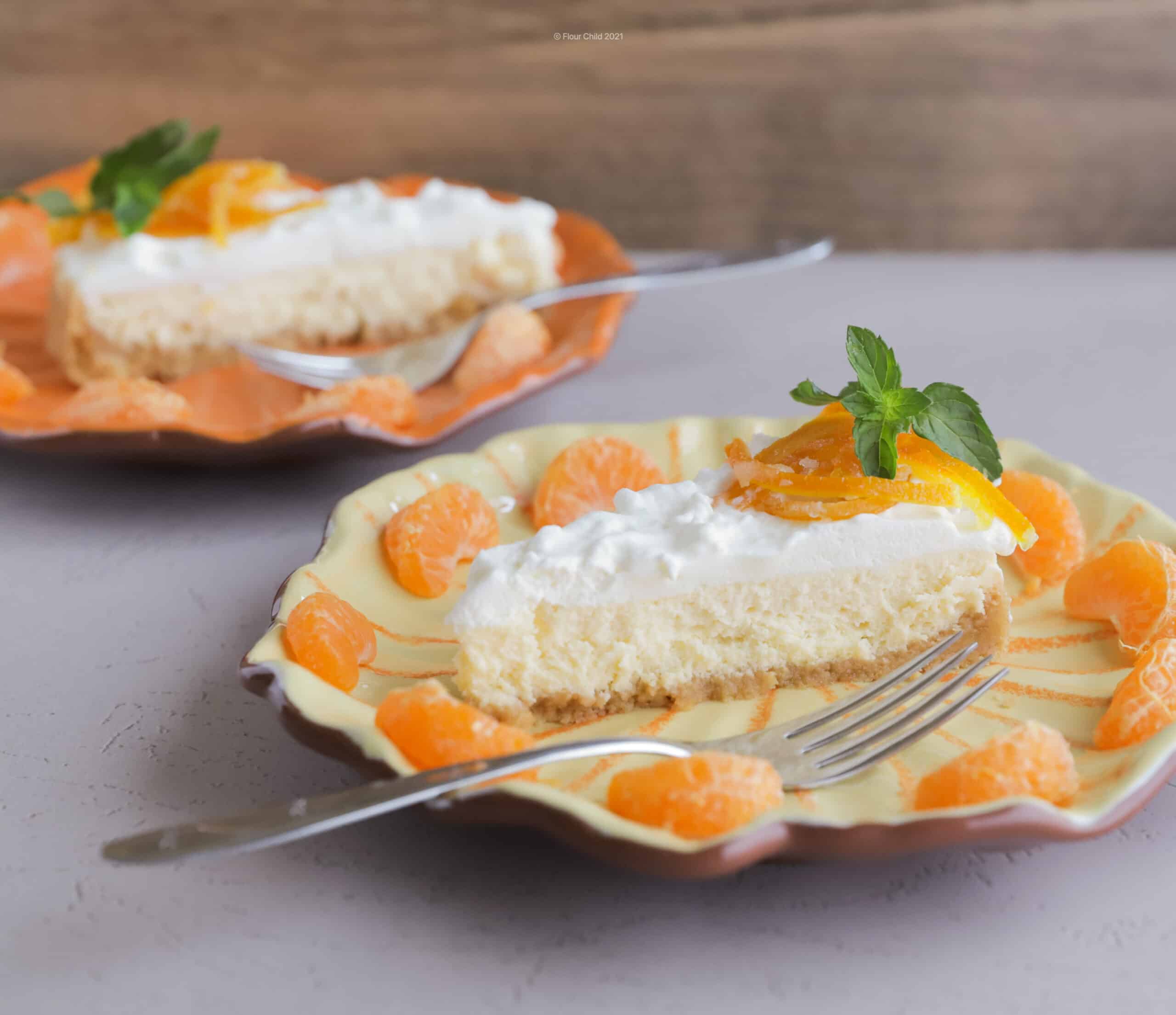 Slice of orange Creamsicle cheesecake on a dessert plate with a fork, garnished with small orange wedges all around. A second plate is in the background. 