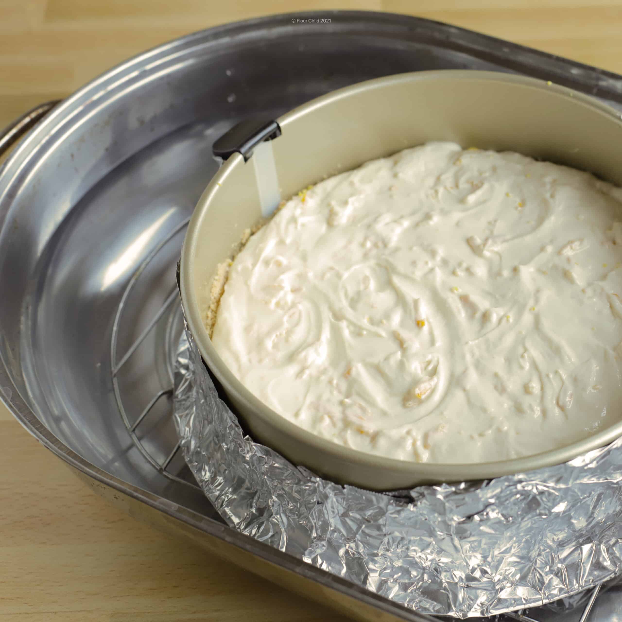 Springform pan with cake batter and wrapped in foil sitting inside a large roasting pan on a rack with water coming a ½ inch up the side of the springform pan