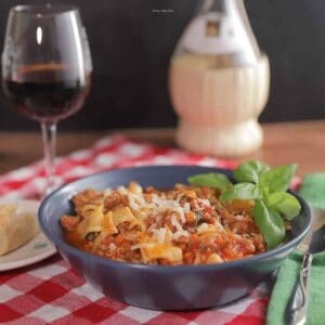 A bowl of lasagna soup with shredded cheese on top and basil garnish. A glass of wine and a hot roll are pictured next to the bowl.