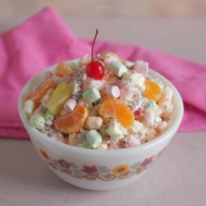 Ambrosia salad with colored marshmallows inside an antique bowl with a pink napkin behind it.