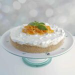 Orange creamsicle cheesecake on a cake pedestal with candied orange peel and mint leaves garnishing the top in the center.