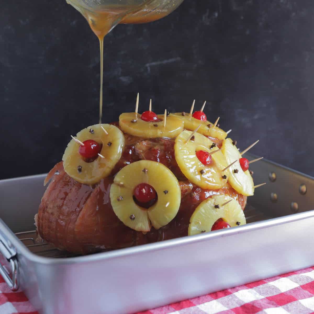 A spiral sliced ham with pineapple and cherries affixed with toothpick, sitting in a large silver roasting pan.