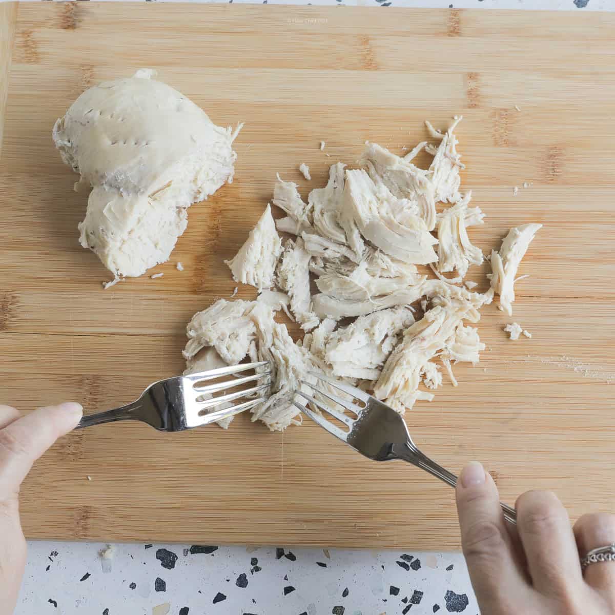 Cooked chicken breast on a cutting board, being pulled apart using two forks.