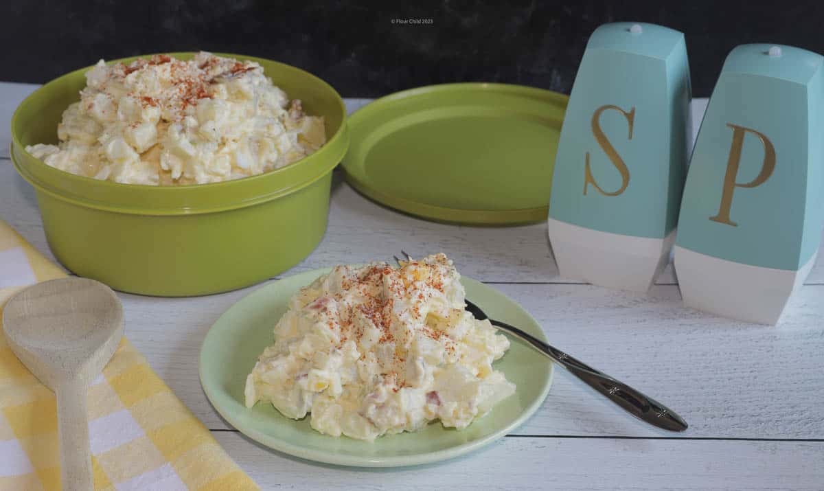 A serving of potato salad on a small green salad plate, with paprika sprinkled on top. Salt shaker and serving bowl of potato salad sitting next to plate. 