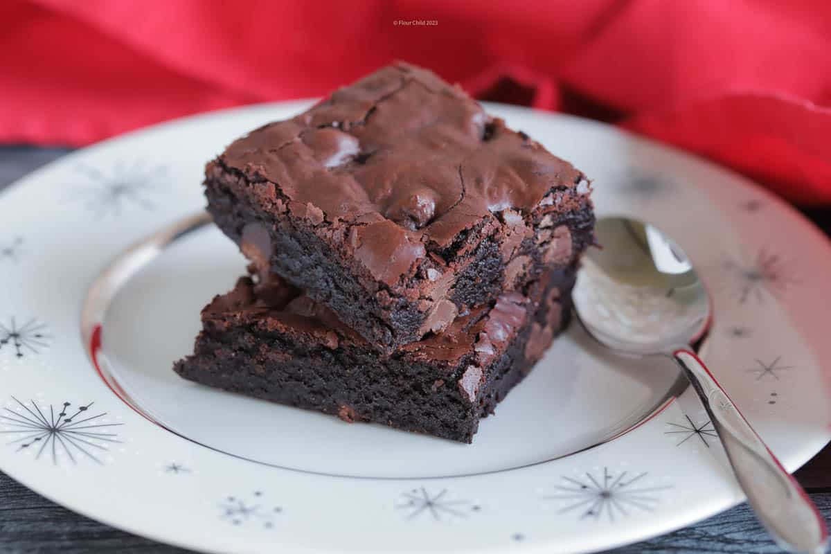 Two dark chocolate brownies stacked on top of each other on an antique white and silver dessert plate.