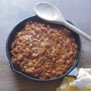 A cast iron skillet full of barbecued beans, with a wooden spoon set on the edge of the pan.