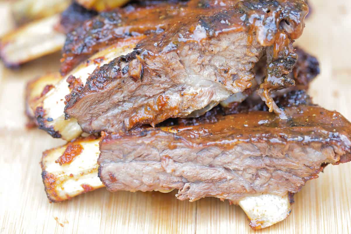 A stack of grilled beef ribs basted with barbecue sauce on a wooden cutting board.